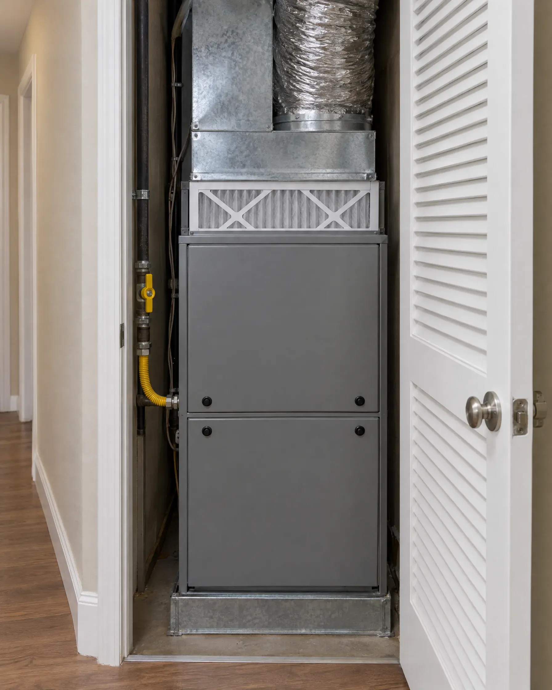 A gas furnace in a hallway utility closet with the louvered door open, a fresh filter in its slot and a yellow gas shutoff valve.